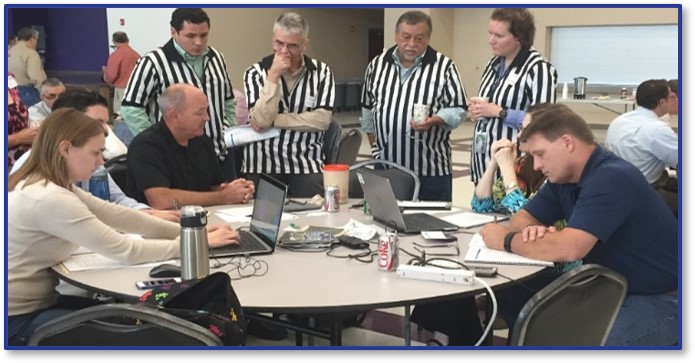 Photo of a multihazard tournament, in which four individuals in referee uniforms stand over and discuss with individuals working on computers at a table